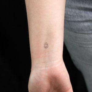 Puede incluir: Un pequeño tatuaje de línea negra de una cara sonriente con cuernos de diablo y un halo en la muñeca de una persona.