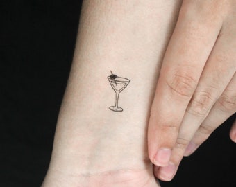 Op de afbeelding: Een kleine zwarte lijntekening van een martini-glas met een olijf op een stokje. Het glas is gevuld met vloeistof. De tekening bevindt zich op de pols van een persoon.