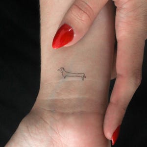 Puede incluir: Un pequeño tatuaje minimalista de línea de un dachshund en la muñeca de una persona.