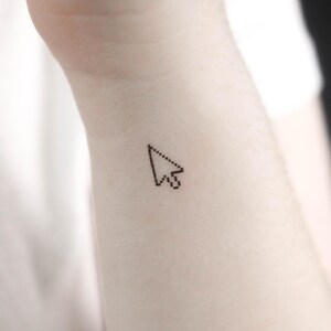 May include: A small black pixelated mouse cursor tattoo on a person's wrist.