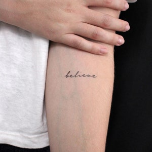 Könnte beinhalten: Eine Nahaufnahme eines Unterarms mit einem einfachen schwarzen Tattoo in Kursivschrift, das "glauben" lautet.