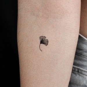 Op de afbeelding: Een eenvoudige zwarte lijntekening van een ginkgo-blad tattoo op iemands arm.