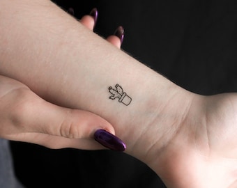 May include: A simple line drawing tattoo of a potted cactus on a person's wrist.