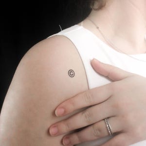 Può includere: Un piccolo tatuaggio nero con il simbolo del copyright sulla spalla di una persona.