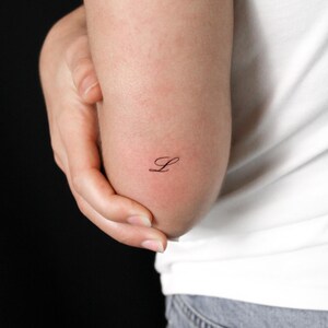 May include: A small, black, fine-line temporary tattoo featuring the cursive letter L. The design is applied to the skin near the elbow, showcasing a delicate, elegant script style with thin, precise strokes that create a clean and minimalist look.