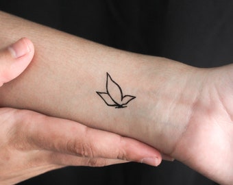 May include: A black temporary tattoo of a stylized leaf design is displayed on a wrist. The tattoo is a simple outline, featuring three connected leaf shapes. The skin tone is light against a dark background.