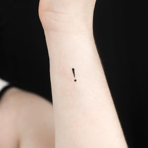 Könnte beinhalten: Nahaufnahme eines Handgelenks mit einem kleinen schwarzen Ausrufezeichen-Tattoo.
