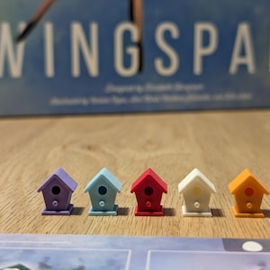 May include: Six colorful birdhouse-shaped game pieces for the board game Wingspan. The pieces are purple, blue, red, white, and orange.