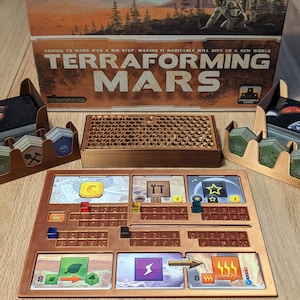 May include: Terraforming Mars board game with a brown wooden game board, a brown wooden tile tray, and a brown wooden card holder. The game board has a green plant icon, a lightning bolt icon, and a temperature icon. The game board also has a star icon and a number 3.