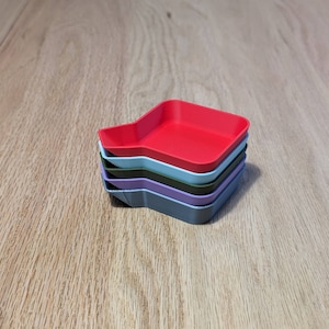 May include: A stack of five small, square trays in different colors. The top tray is red, the next is light blue, the next is green, the next is purple, and the bottom tray is gray.