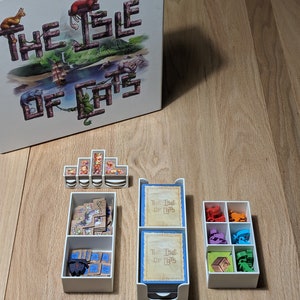 May include: A white box with the game title "The Isle of Cats" printed on the top. The box is open and shows the game components, including wooden tiles, cards, and plastic storage containers.