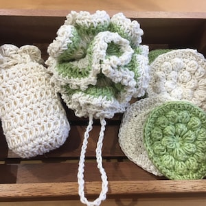 May include: A set of crocheted bath accessories in white and green. The set includes a pouf, a washcloth, and two smaller cloths. The items are displayed in a wooden crate.