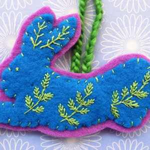 Op de afbeelding: Een blauwe vilt konijn ornament met groene geborduurde bladeren, hangend aan een groene garen lus.