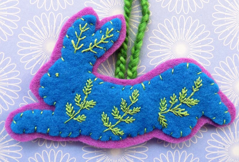 Op de afbeelding: Een blauwe vilt konijn ornament met groene geborduurde bladeren, hangend aan een groene garen lus.