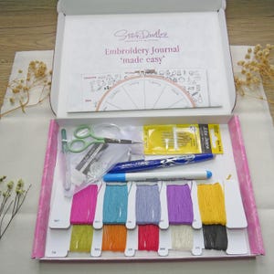 Può includere: Una scatola artigianale aperta con forniture per ricamo. La scatola contiene rocchetti di filo colorati, forbici, aghi, penne e un calendario. La scatola è etichettata "Embroidery Journal 'made easy'" e "Stitch Doodles".