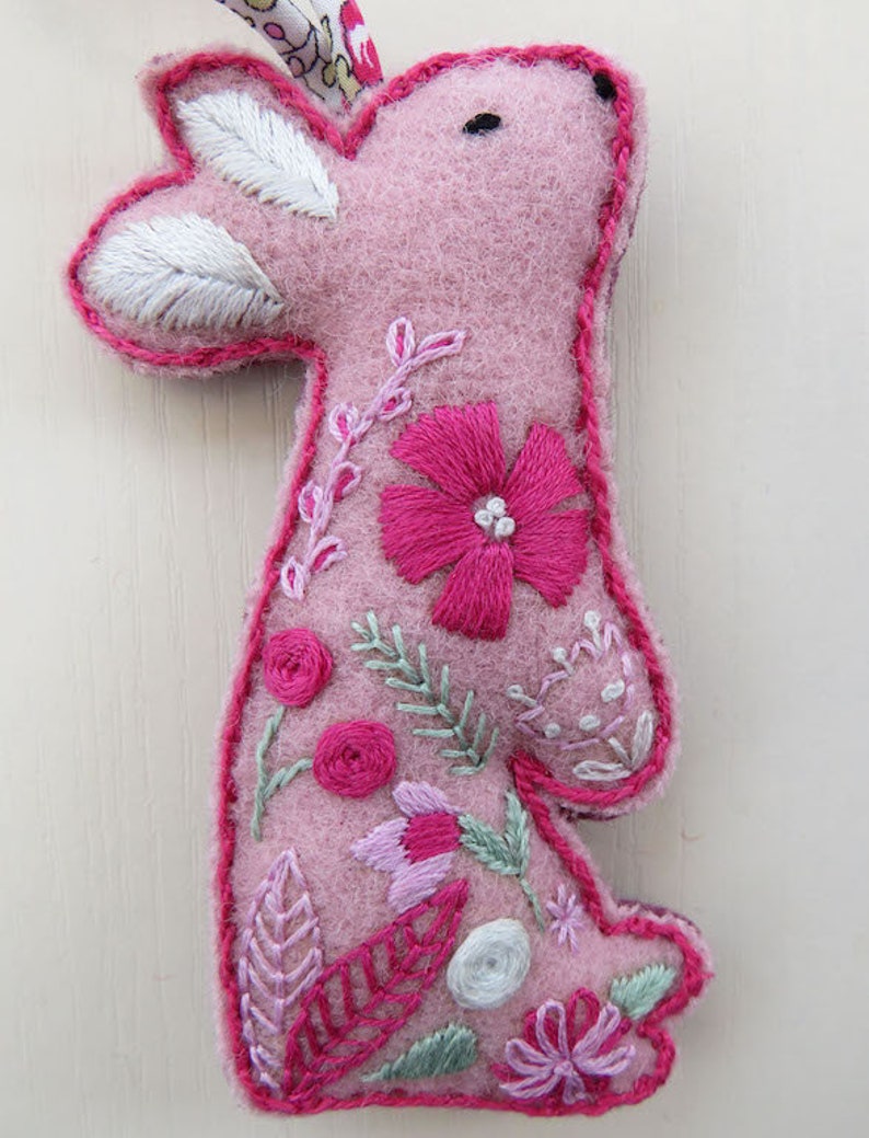 Op de afbeelding: Een roze vilt konijn met geborduurde bloemen en bladeren. Het konijn staat rechtop en heeft een wit vilt oor.