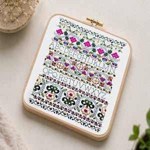 May include: A rectangular embroidery hoop featuring a floral cross-stitch design. The design includes colourful flowers, leaves, and the alphabet in a decorative font. The embroidery is framed in a light-coloured wooden hoop.