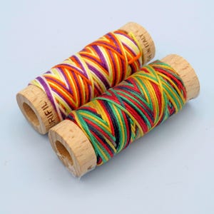 May include: Two spools of colourful embroidery thread, one with orange, yellow, white, and purple thread, and the other with red, yellow, green, black, and blue thread. Both spools are made of wood and have the brand name "URIFIL" printed on them.