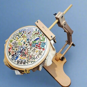 May include: A wooden embroidery hoop stand with a circular hoop holding a colorful embroidered design of flowers, birds, and butterflies. The stand is made of natural wood and has a simple, elegant design. The hoop is attached to the stand with a metal clamp. The stand is labeled "Nudge".