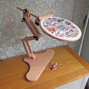 Puede incluir: Un soporte de bordado de madera con un aro redondo que sostiene un diseño de punto de cruz colorido. El soporte es ajustable y descansa sobre una base plana. Dos carretes de hilo están cerca. El soporte es de madera clara.