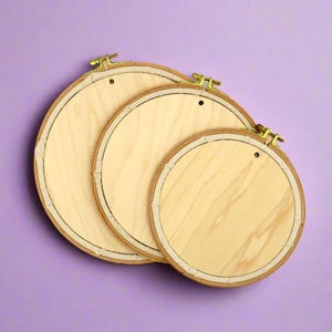 May include: Three wooden embroidery hoops of different sizes, with a light brown wood colour and a natural finish. The hoops are nested together, with the largest hoop at the bottom and the smallest hoop at the top. The hoops are on a purple background.