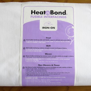 Könnte beinhalten: Eine weiße Rolle HeatnBond Bügeleinlage mit einem lila Etikett. Das Etikett zeigt den Markennamen, die Produktart und die Bügelanleitung. Der Text auf dem Etikett beschreibt verschiedene Arten von Einlagen: Tricot, Weft, Woven und Non-Wovens & Fleece.