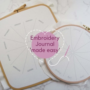 May include: Two embroidery hoops with a white fabric stretched over them. The fabric is divided into 12 sections, each labeled with a month of the year. The text "Embroidery Journal 'made easy'" is written in a pink heart shape on top of the hoops.
