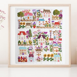 Könnte beinhalten: Ein farbenfrohes gesticktes Design einer kleinen Stadt mit Häusern, Bäumen und Blumen. Die Häuser sind weiß mit roten, rosa und blauen Akzenten. Die Bäume sind grün und die Blumen sind rosa, lila und gelb.