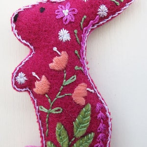 Op de afbeelding: Een roze vilt konijn met geborduurde bloemen en bladeren. Het konijn staat op een witte achtergrond.