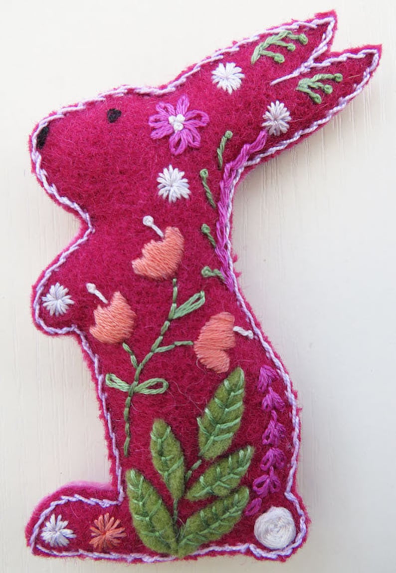 Op de afbeelding: Een roze vilt konijn met geborduurde bloemen en bladeren. Het konijn staat op een witte achtergrond.