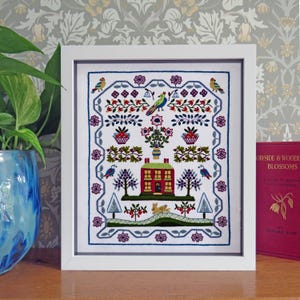May include: A framed embroidery sampler featuring a central red house with a green roof, surrounded by colorful floral motifs, trees, birds, and a small rabbit. The design is enclosed within a decorative blue border on a white fabric background.