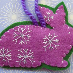 Op de afbeelding: Een paarse vilt konijn ornament met witte geborduurde sneeuwvlokken. Het konijn is omrand met groen vilt en heeft een paarse garen lus om op te hangen.