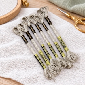 May include: Five skeins of metallic silver embroidery floss arranged in a fan shape. Each skein features a black and yellow paper label with the text E168 and a barcode. The thread has a shimmering, reflective texture suitable for needlework.