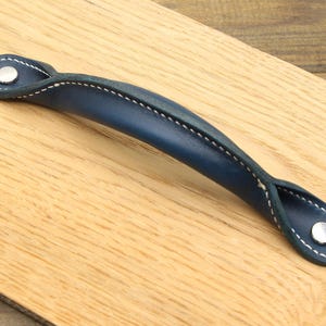 May include: A navy blue leather handle with white stitching and silver rivets. The handle is curved and appears to be attached to a wooden surface. The handle is a decorative element for furniture.