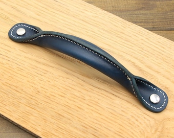 Puede incluir: Un tirador de cuero azul marino con costuras blancas y remaches plateados. El tirador es curvo y está fijado a una superficie de madera. Es un elemento decorativo para muebles.