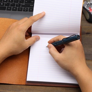 May include: A person writing in a brown leather-bound journal with a blue pen. The journal is open to a page with lined paper. A computer keyboard and a mouse are visible in the background.