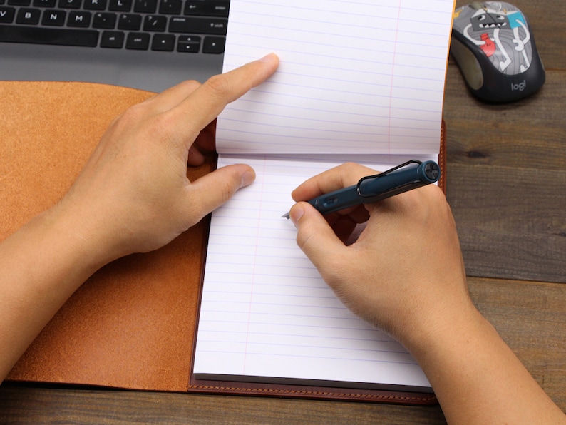 May include: A person writing in a brown leather-bound journal with a blue pen. The journal is open to a page with lined paper. A computer keyboard and a mouse are visible in the background.