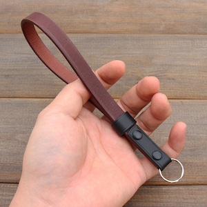 Pu&ograve; includere: Un cinturino per fotocamera in pelle marrone con un anello metallico nero e due bottoni neri.