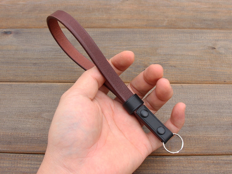 Pu&ograve; includere: Un cinturino per fotocamera in pelle marrone con un anello metallico nero e due bottoni neri.