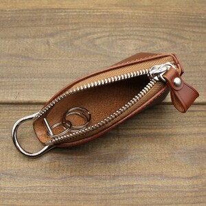 May include: A brown leather key holder with a zipper closure and a silver metal ring. The key holder is open and two silver metal keys are inside.