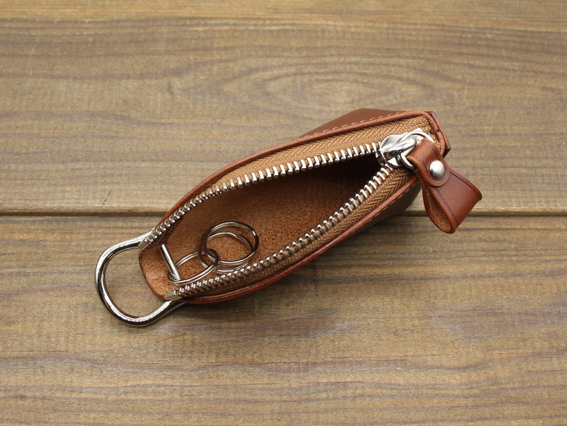 May include: A brown leather key holder with a zipper closure and a silver metal ring. The key holder is open and two silver metal keys are inside.