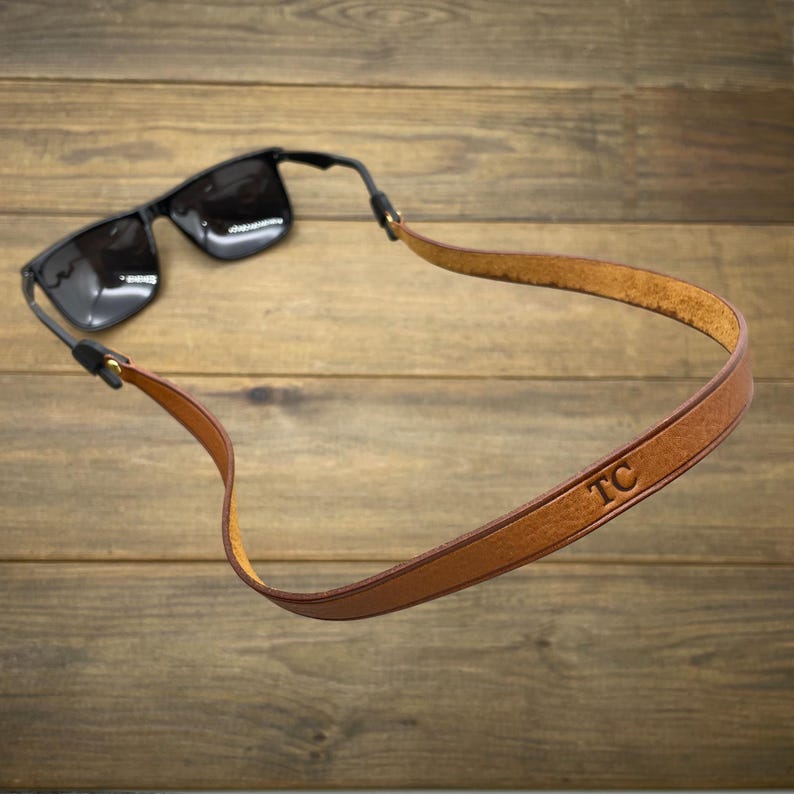 May include: Black sunglasses with a brown leather strap. The strap has the initials "TC" embossed on it. The sunglasses and strap are set against a wooden background.