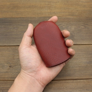 Puede incluir: Una bolsa de cuero roja con una superficie lisa y texturizada. La bolsa est&aacute; cerrada y tiene una forma redondeada.