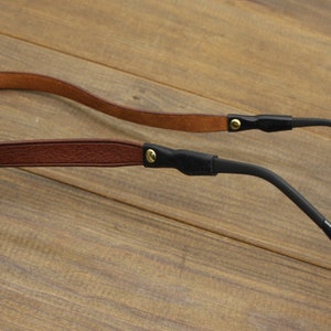 May include: A close-up of a pair of eyeglasses with a brown leather strap and black temples. The strap is attached to the temples with small gold-colored rivets. The eyeglasses are set against a wooden background.