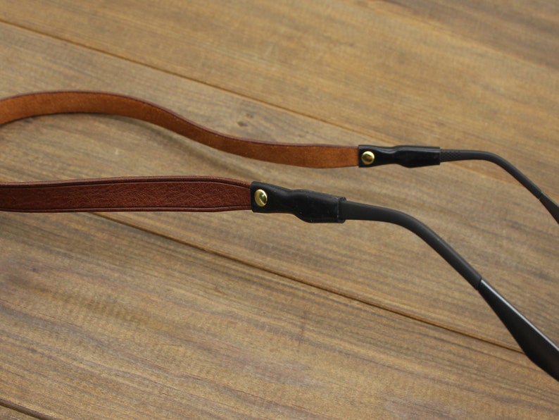 May include: A close-up of a pair of eyeglasses with a brown leather strap and black temples. The strap is attached to the temples with small gold-colored rivets. The eyeglasses are set against a wooden background.