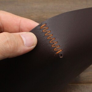 May include: Close-up of a dark brown leather piece with orange stitching. The leather is folded in half and the stitching is visible along the fold.