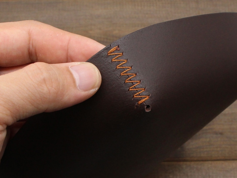 May include: Close-up of a dark brown leather piece with orange stitching. The leather is folded in half and the stitching is visible along the fold.