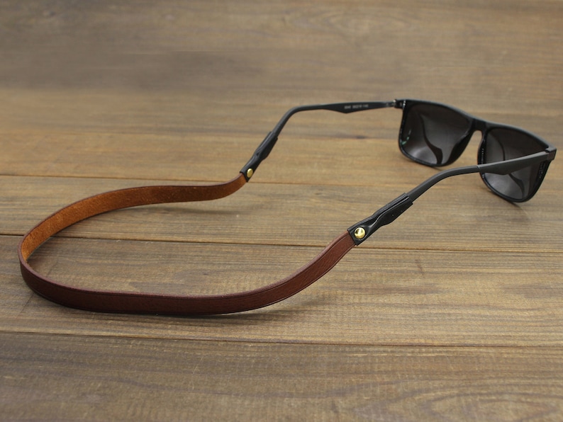 May include: Sunglasses with a brown leather strap and black frames. The strap is attached to the temples with black connectors and gold-colored rivets. The sunglasses are resting on a wooden surface.