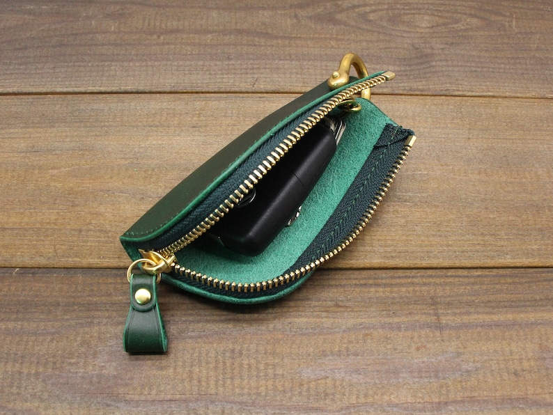 Puede incluir: Un llavero de cuero verde con una cremallera dorada y un anillo dorado. El llavero est&aacute; abierto y hay una llave negra dentro.