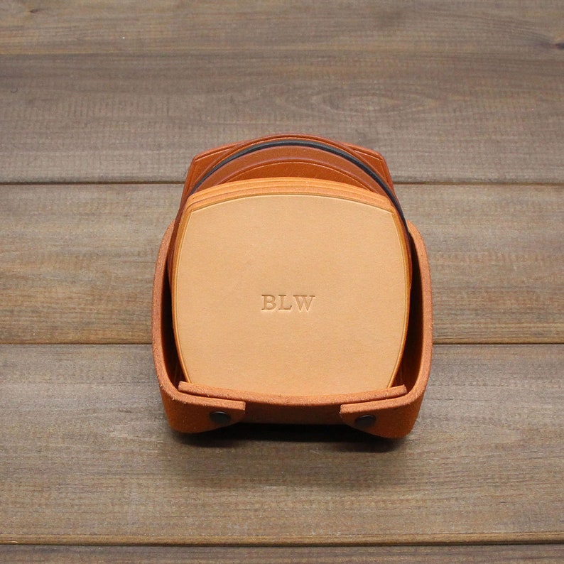 Puede incluir: Un juego de posavasos de cuero marr&oacute;n con un soporte de cuero. Los posavasos est&aacute;n apilados y el posavasos superior tiene las iniciales "BLW" grabadas.